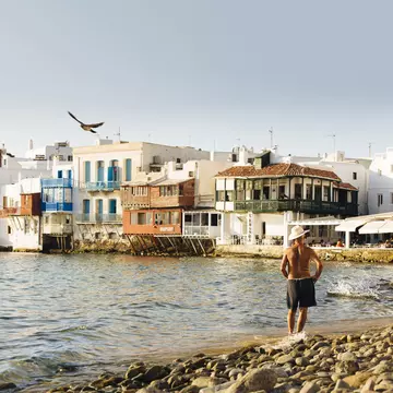 A man in shorts and a hat stands on the shore facing a row of beautiful buildings in Mykonos, Greece, as the evening light begins to fade. 