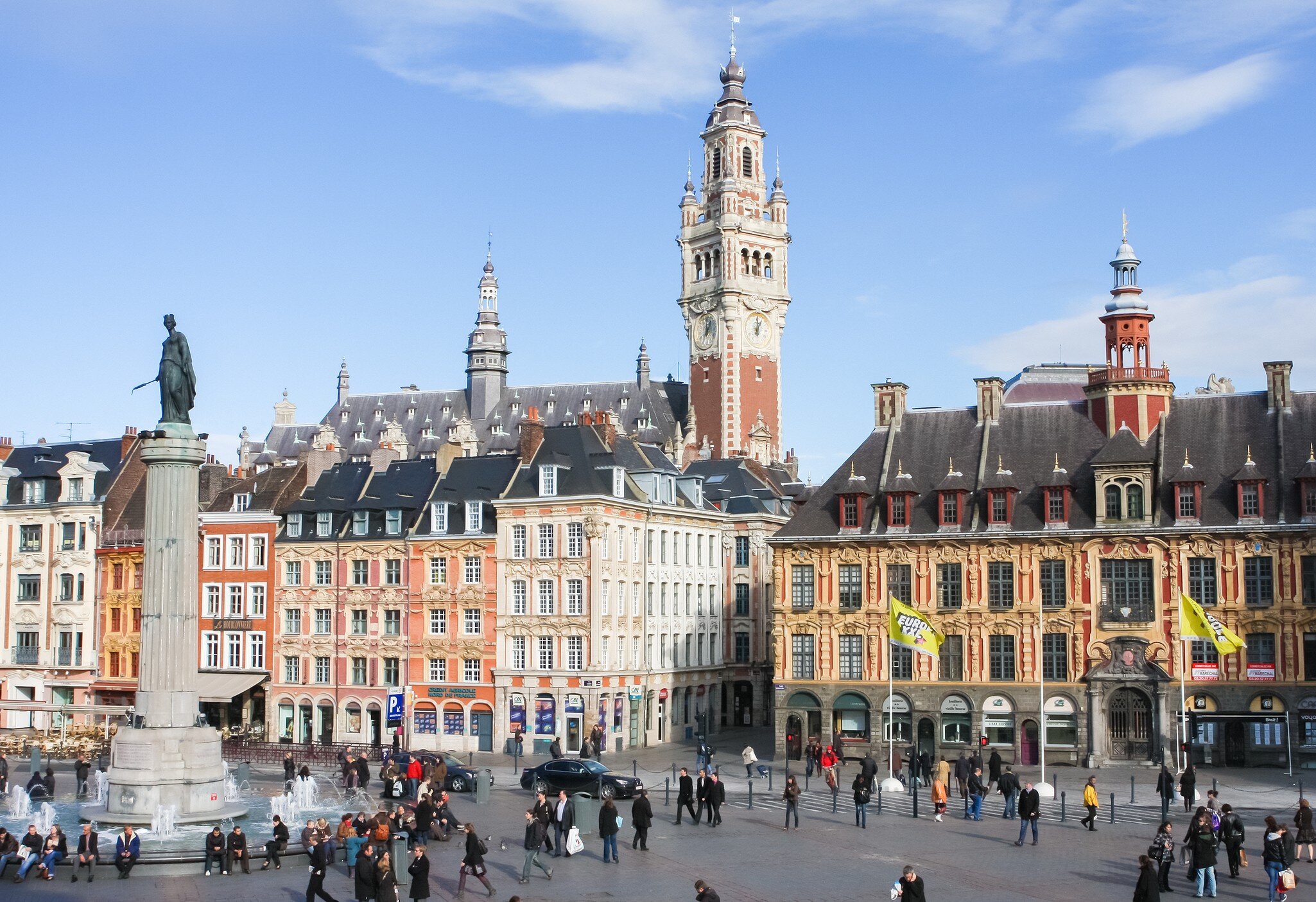 Brick-and-stone clock tower rising behind ornate gabled facades, with a column statue, fountain and crowds on a square