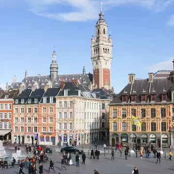 Brick-and-stone clock tower rising behind ornate gabled facades, with a column statue, fountain and crowds on a square