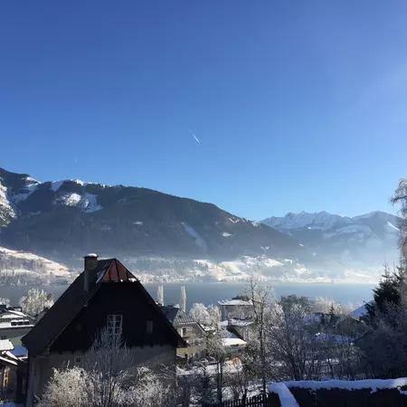 Snow on mountains with evergreen trees; a peaked roof and bare trees are in the foreground.