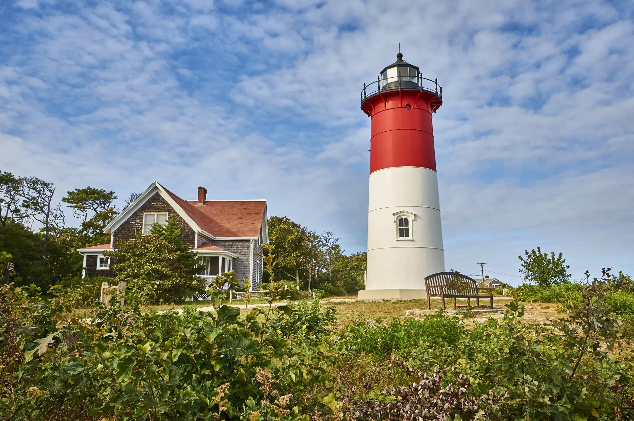 A lighthouse that's half red and half white; a shingled structure is behind it and a bench on the grass in front.