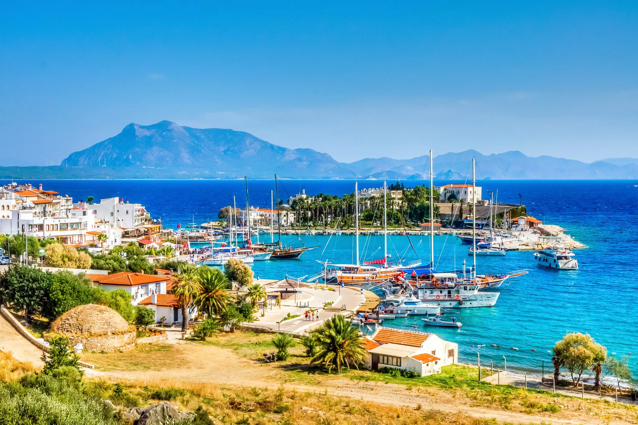 Boats in a harbor of azure water; there is a small town of white buildings on the shore and mountains in the distance.