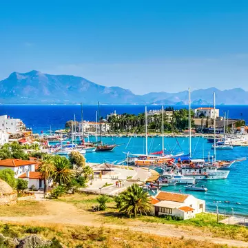 Boats in a harbor of azure water; there is a small town of white buildings on the shore and mountains in the distance.