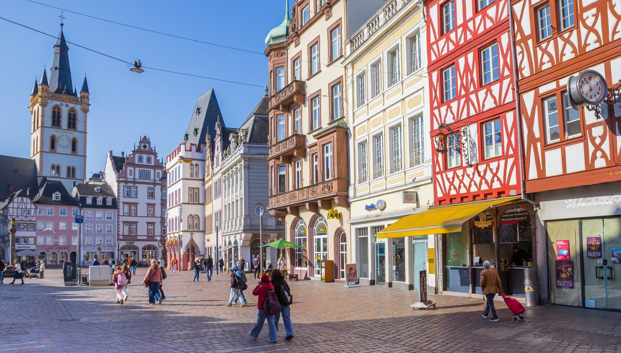 Shoppers on the Hauptmarkt in Trier, Germany, below half-timbered and pastel facades and the tower of St Gangolf