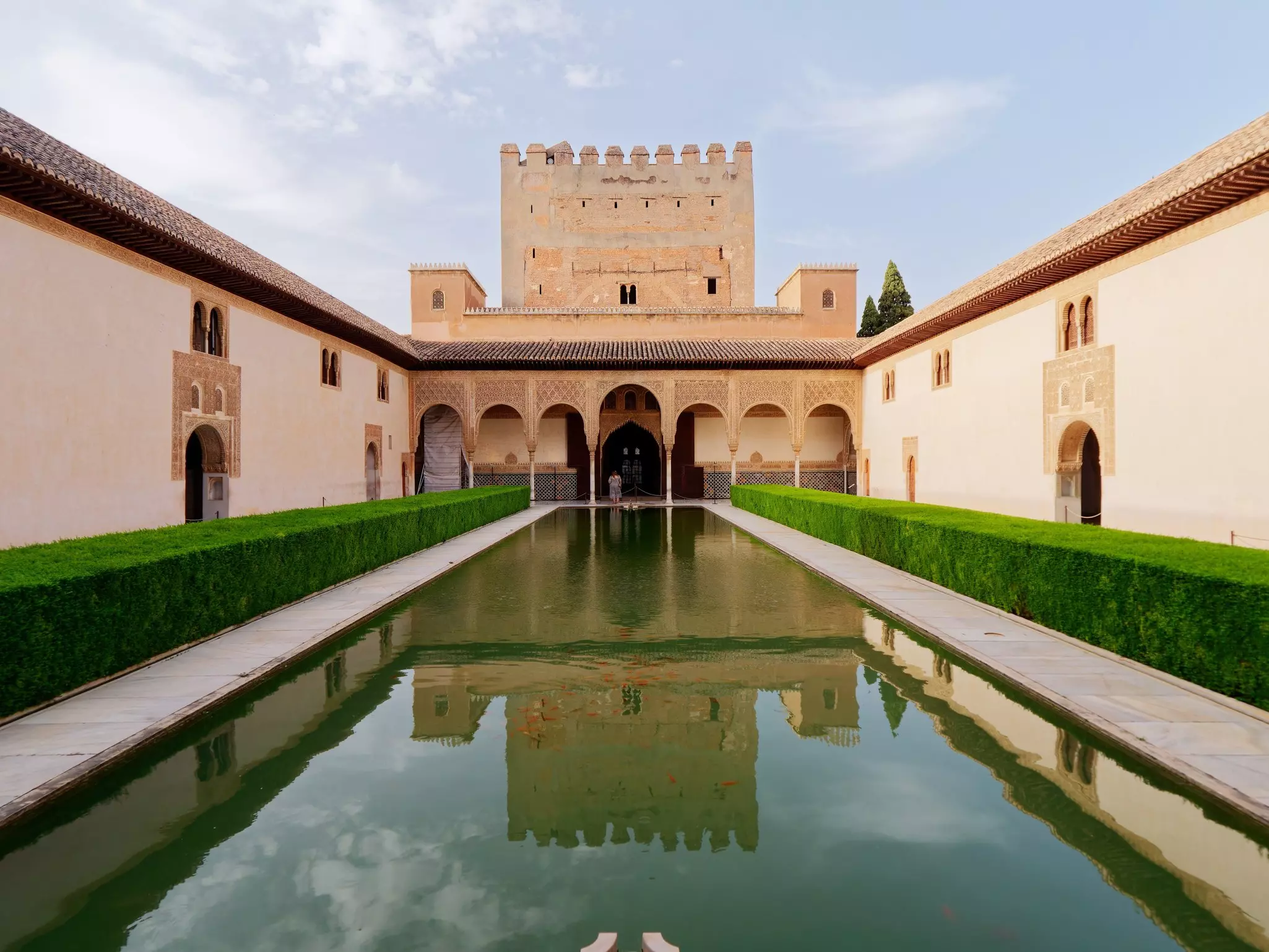 A tan palace is reflected in a pool in a courtyard with green hedges.