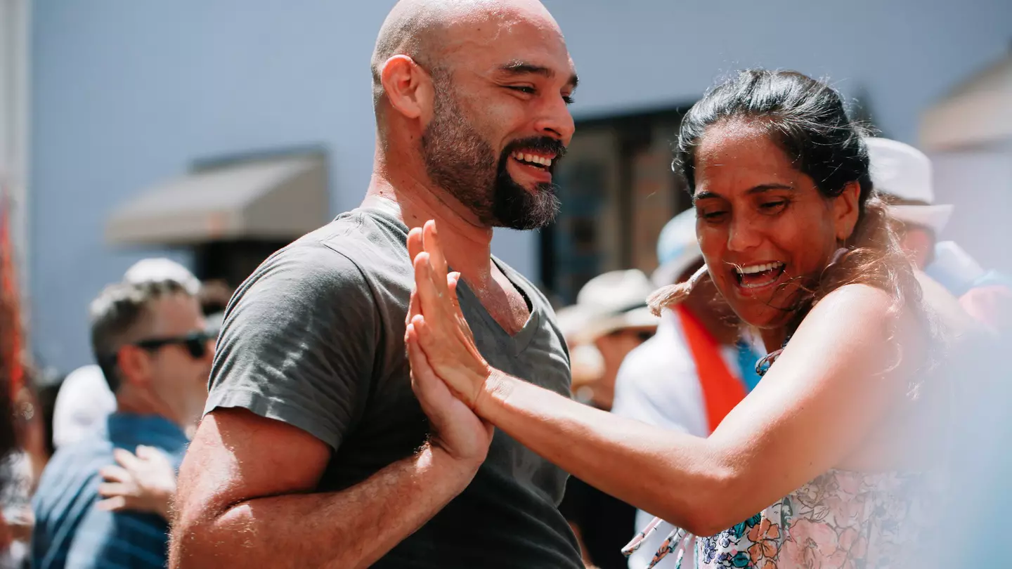 A man and a woman touch hands as they dance to salsa music outside with a group of people.