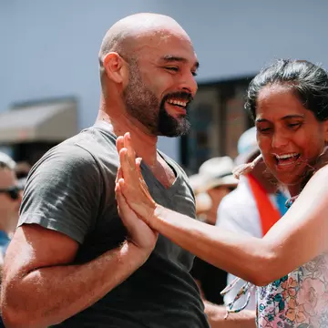 A man and a woman touch hands as they dance to salsa music outside with a group of people.