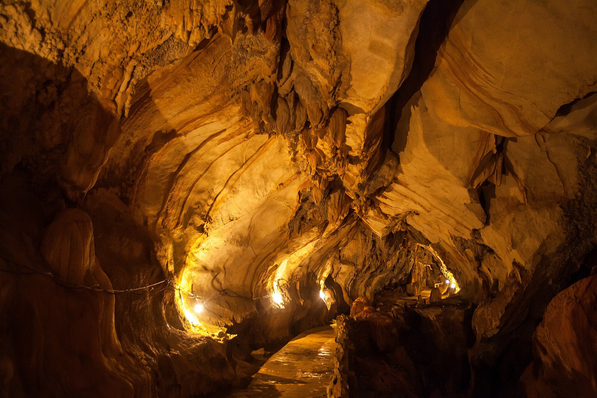 A cave interior of folded, layered rock lit warm gold, with a narrow walkway curving away through it.
