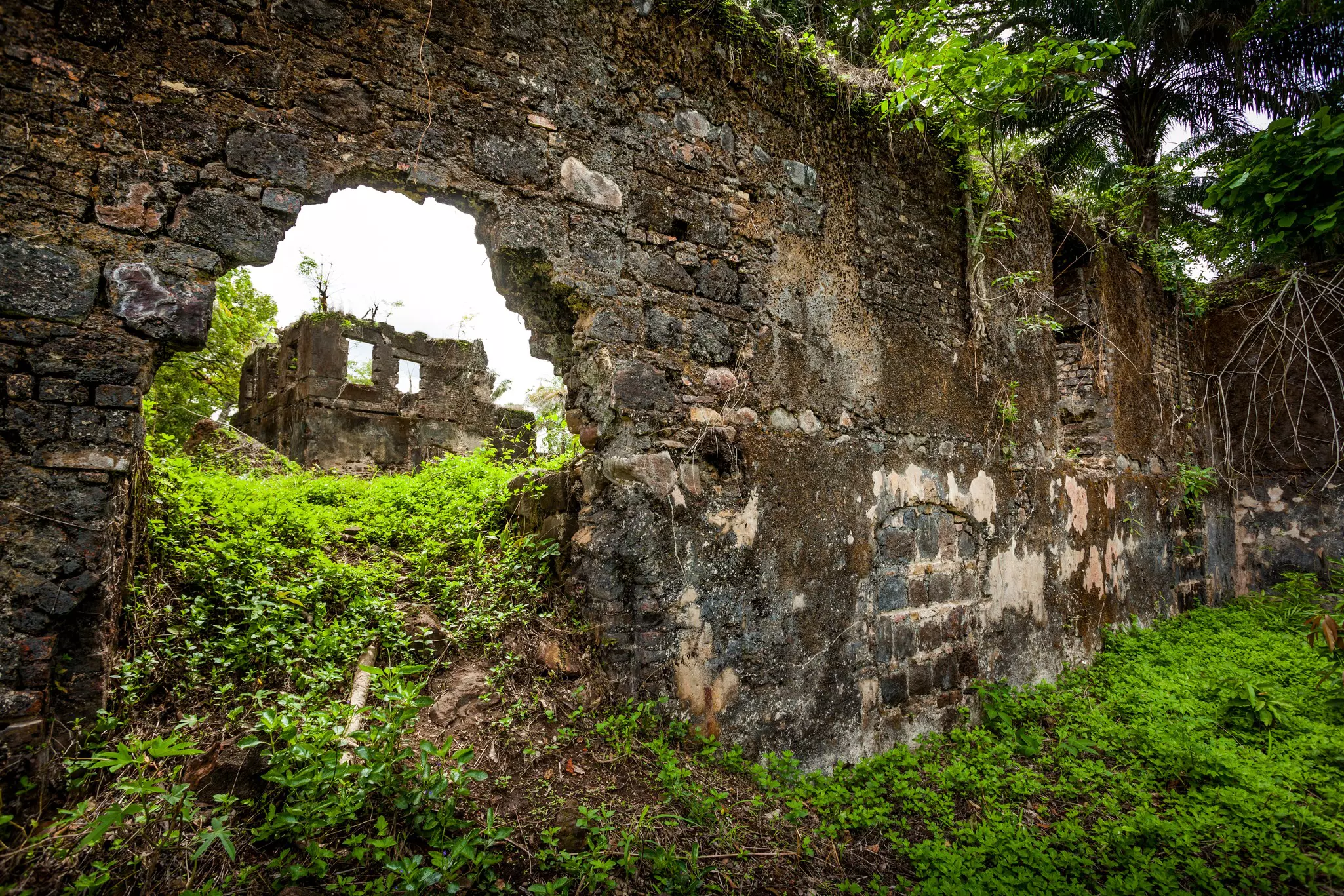 An abandoned, ruined building is seen through a hole in a deteriorating stone wall at a complex that is overgrown with vegetaion.
