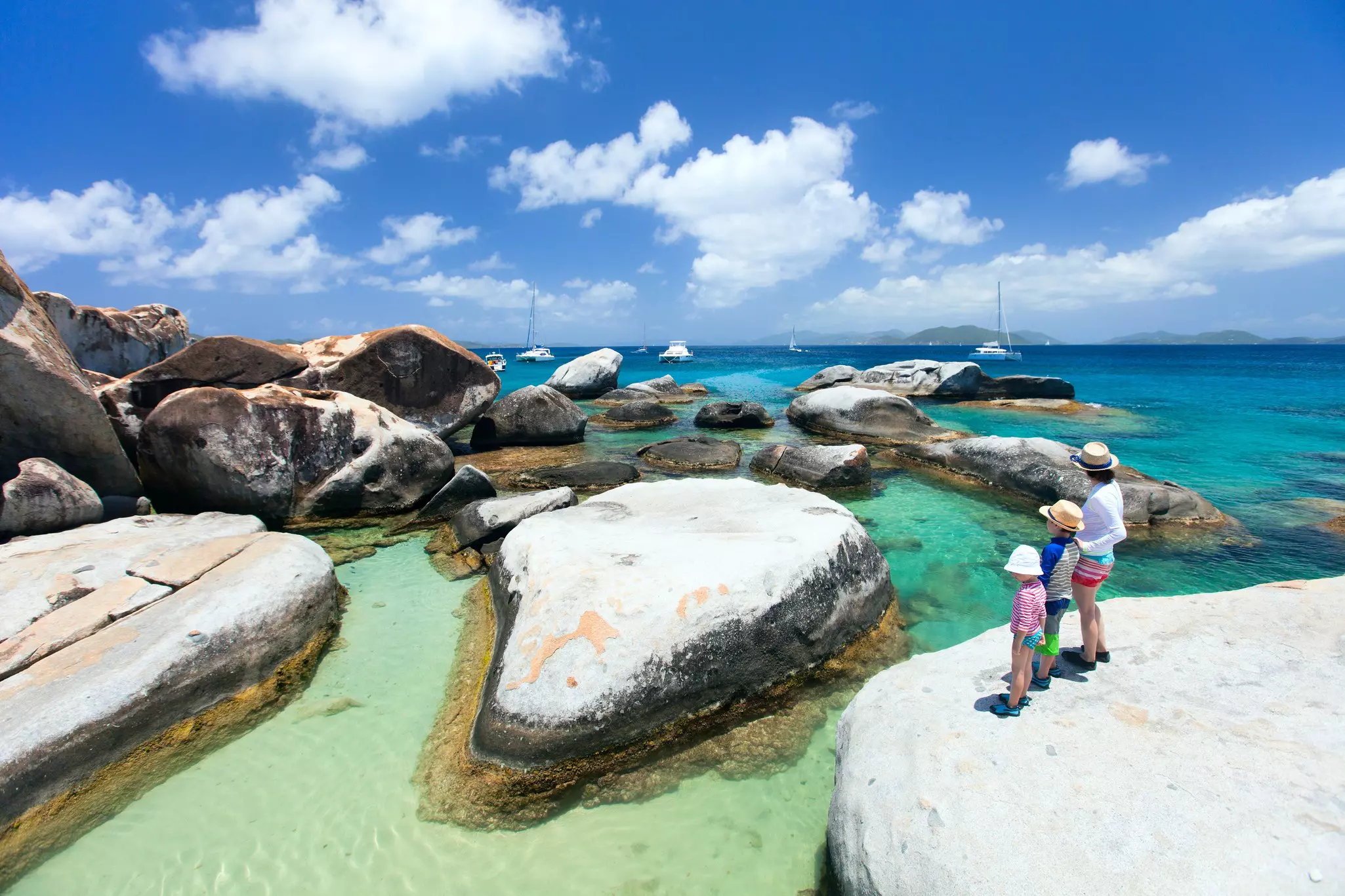A mother and two children stand on a boulder overlooking other rocks by the sea in a tropical spot.