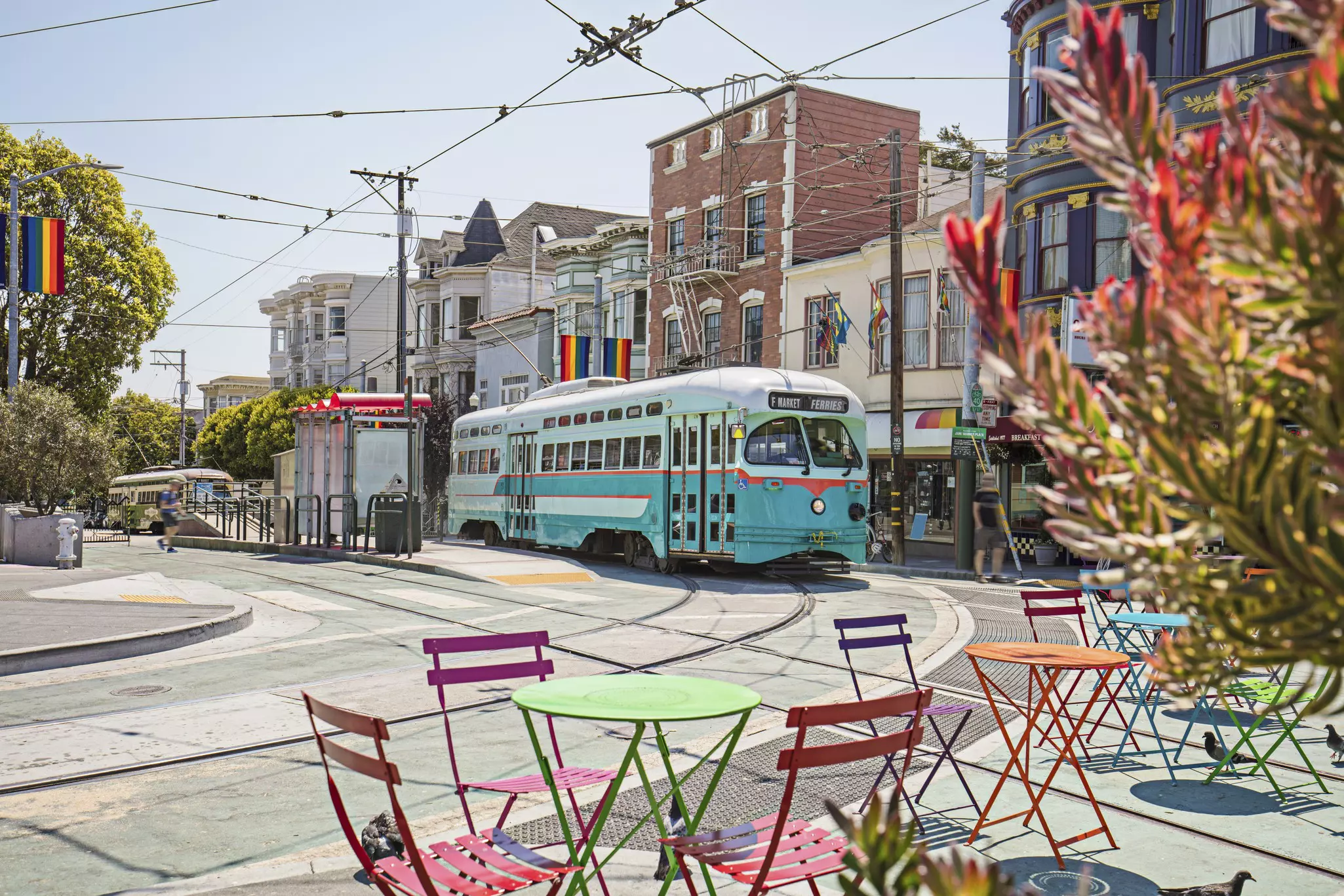 A blue streetcar with rainbow-colored bistro seating in the foreground and rainbow flags along buildings in the background.