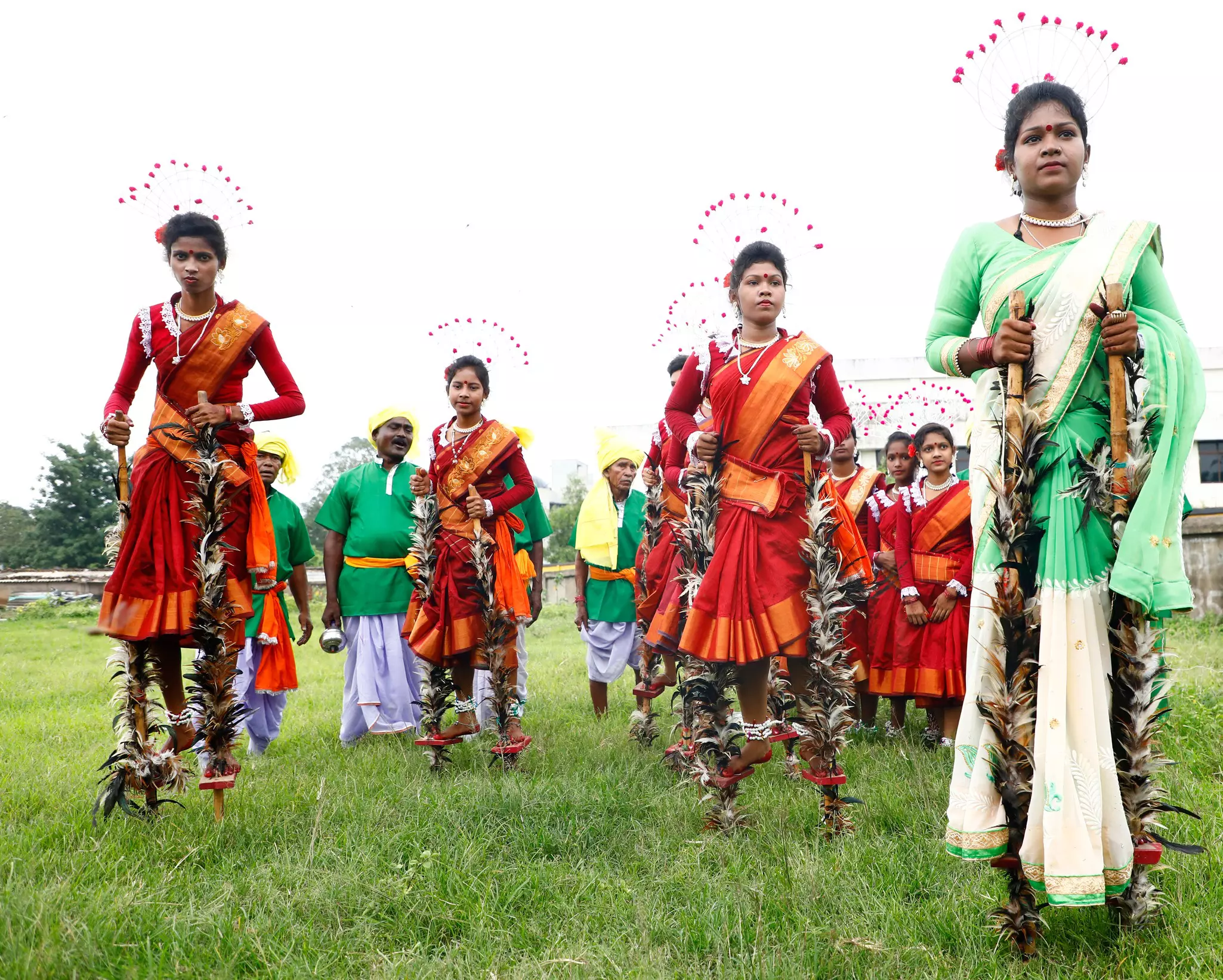 Women in the traditional dress of their tribe - some in red and orange, one in green and white - wear headdresses and stand on low-level stilts with poles covered in feathers.