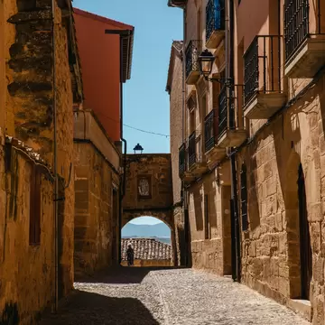 Narrow cobblestone street between golden stone houses with iron balconies, ending in an arch with hills beyond.