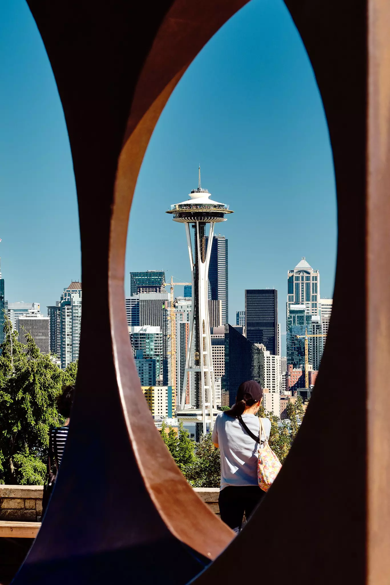 A vertical shot of the Space Needle in Seattle Washington through the framed sides of a curved art installation. A woman is photographing the Space Needle in the foreground.