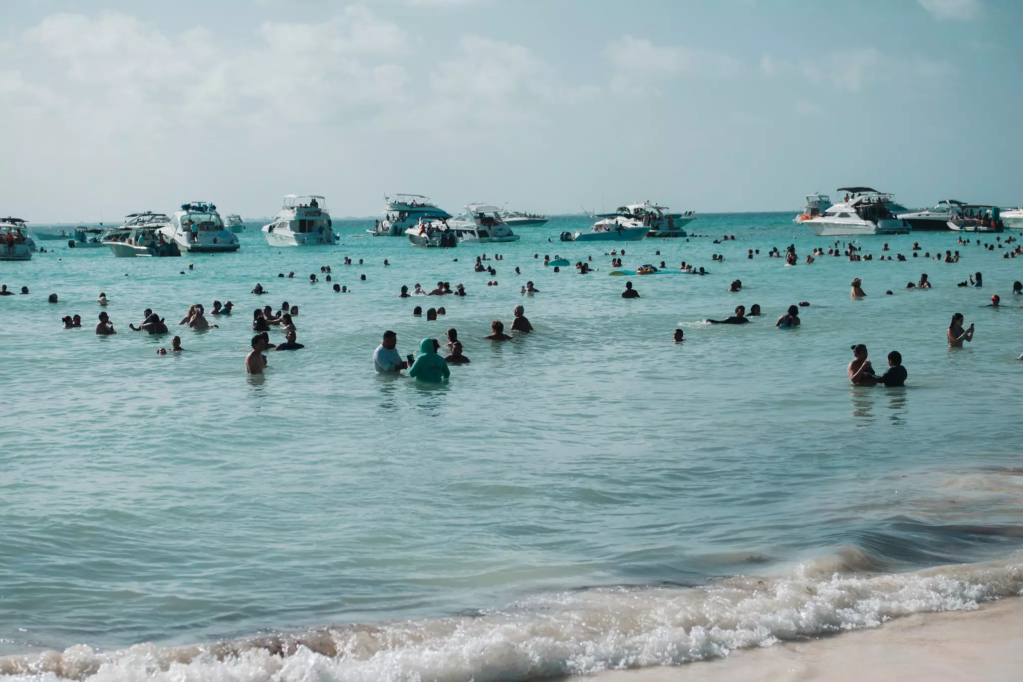 People stand and swim in blue water just off a sandy beach; several boats are stopped offshore.