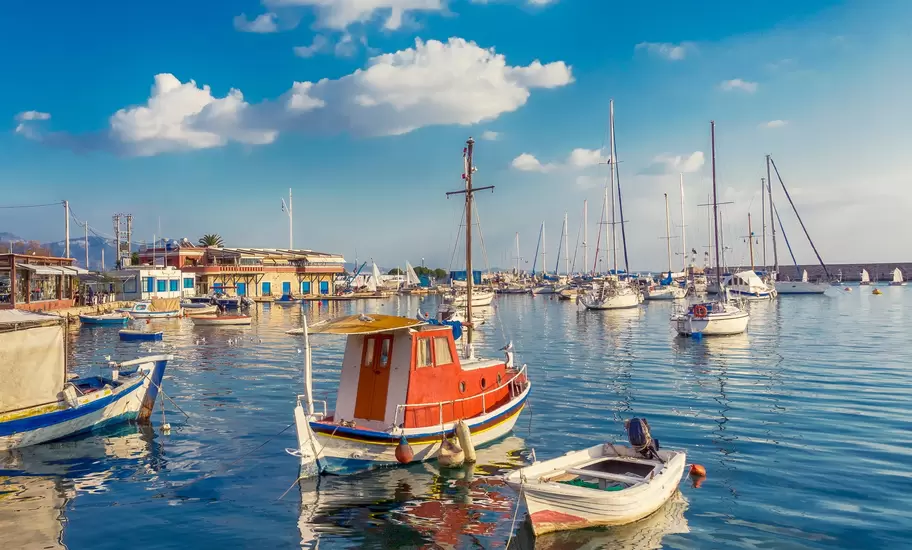 Red and white wooden fishing boat and rowboat moored in a calm Greek harbour with sailing yachts and waterfront buildings