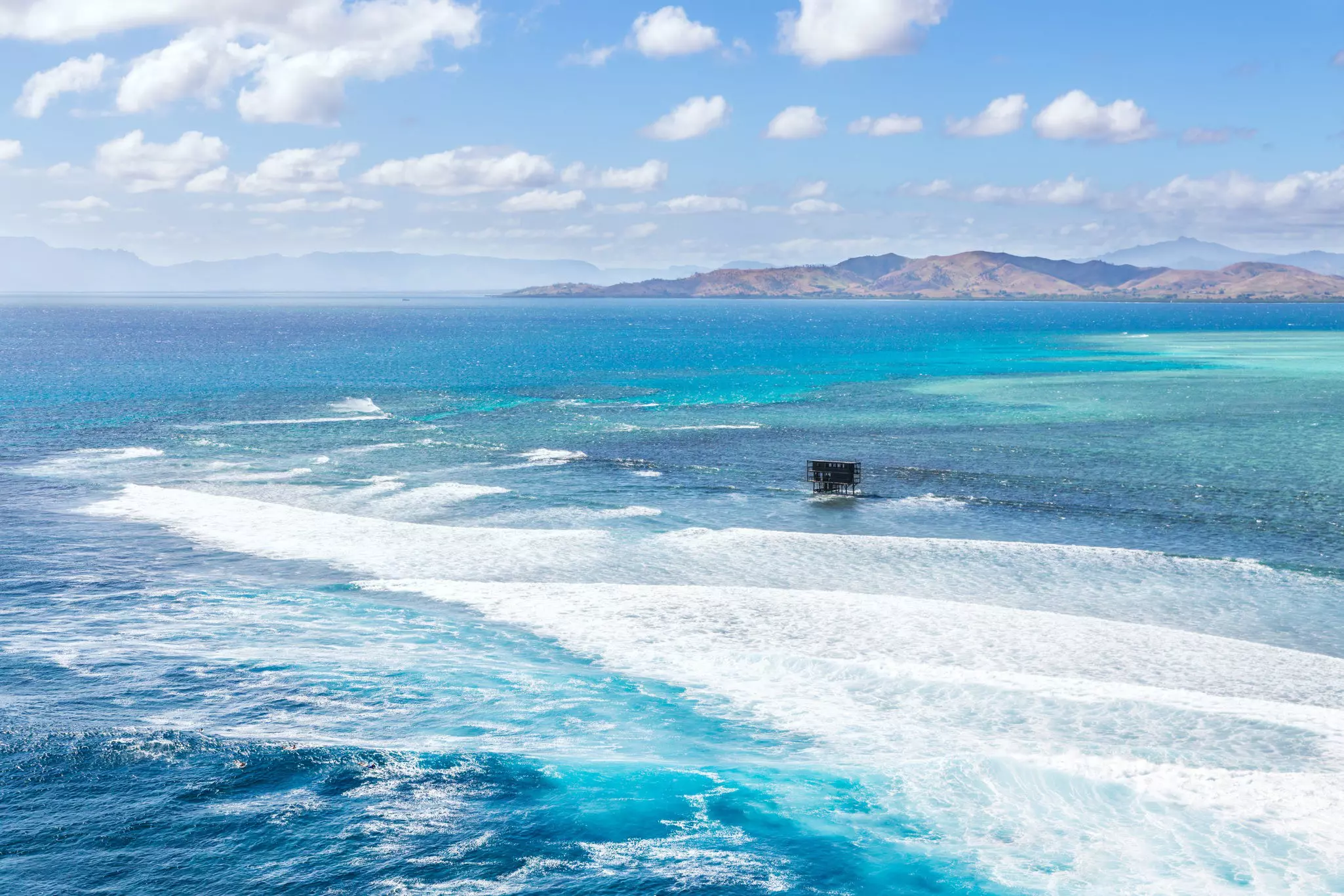 A wooden structure in the ocean stands near a surf break. The surrounding ocean is turquoise, blue and green.