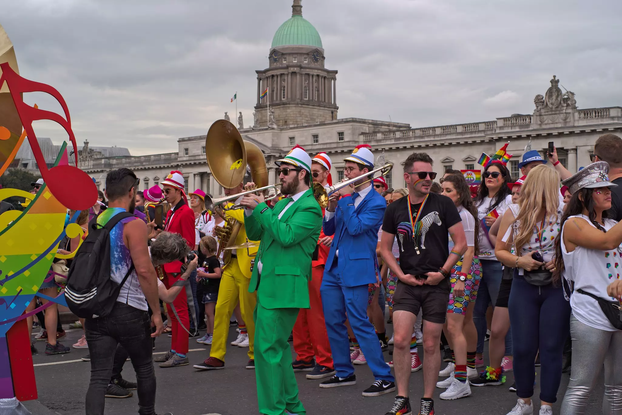 Musicians in colorful costumes play for people on the streets during a festival. An elegant domed building is pictured in the background.