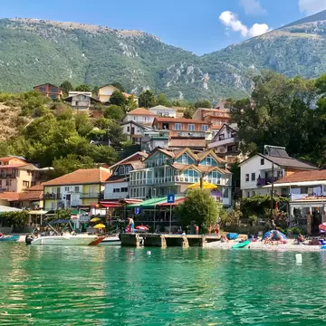 A small lakeside village backed by mountains with people relaxing on the sandy beach