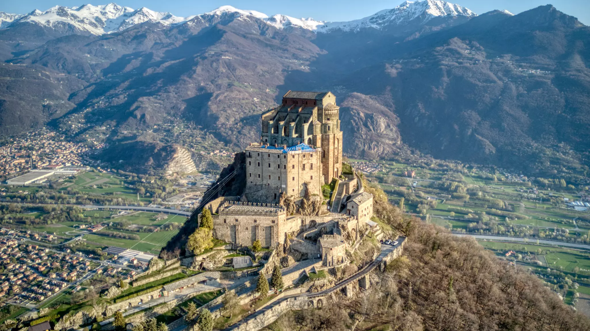 An aerial view of a fortified hilltop monastery complex. A green valley lies below, and snow-capped peaks can be seen in the distance.