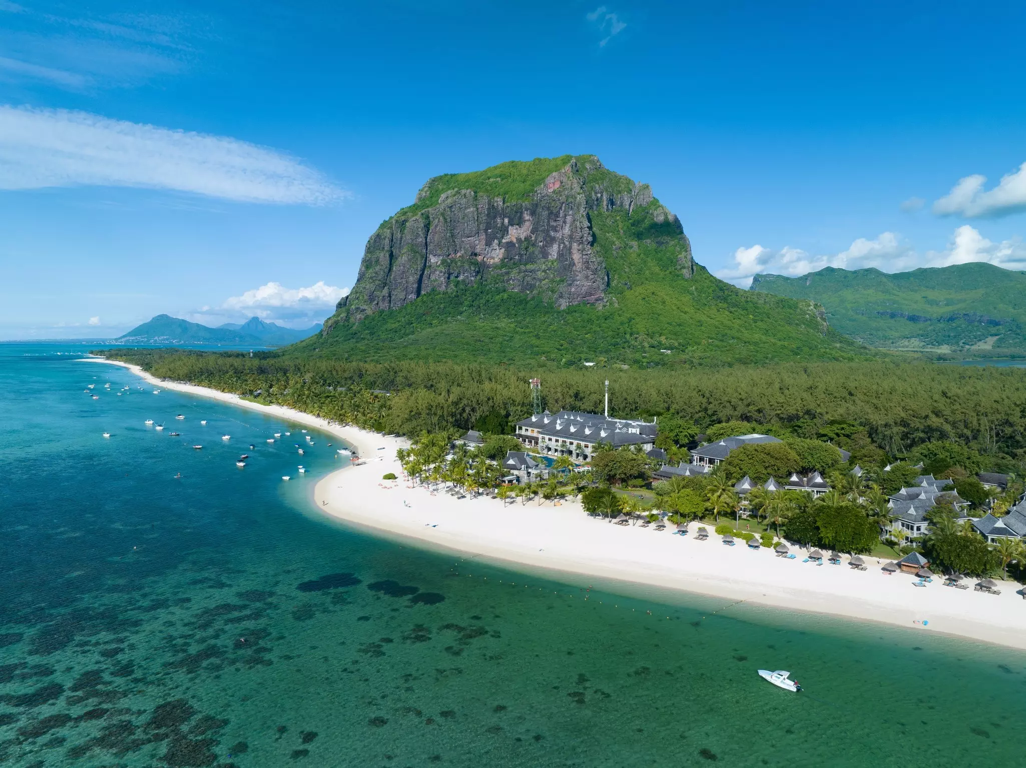 An aerial view of a luxury tropical beach resort, with a dramatic mountain behind and sailboats moored in the shallow water just offshore.
