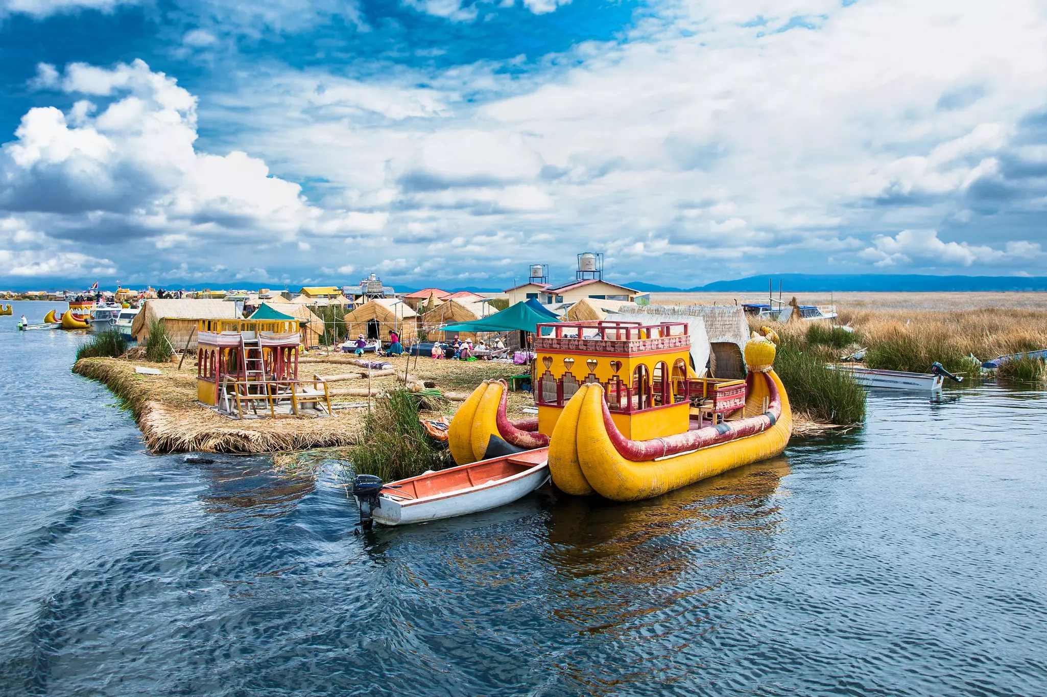 A wide view of traditional boats moored by an island in a lake. Small houses are seen just inland from the shoreline.