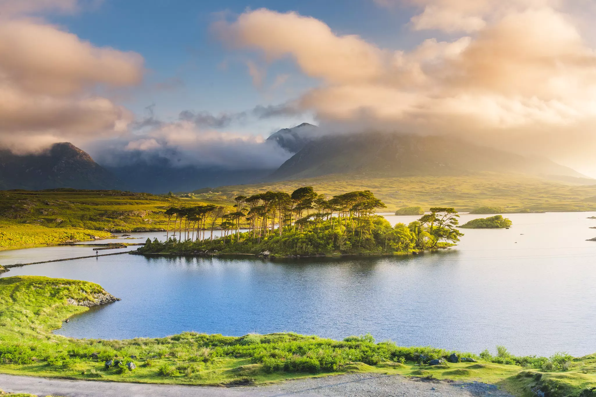 A tree-covered islet in a still lake. Misty clouds cover the top of the surrounding mountains.