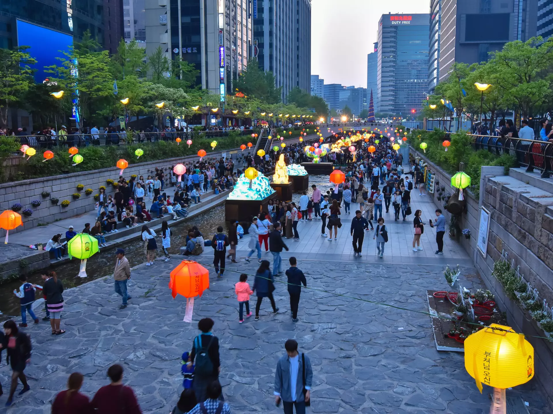 Paper lanterns hang over a public park and promenade in the built-up central business district of a city.