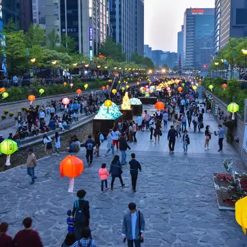 Paper lanterns hang over a public park and promenade in the built-up central business district of a city.