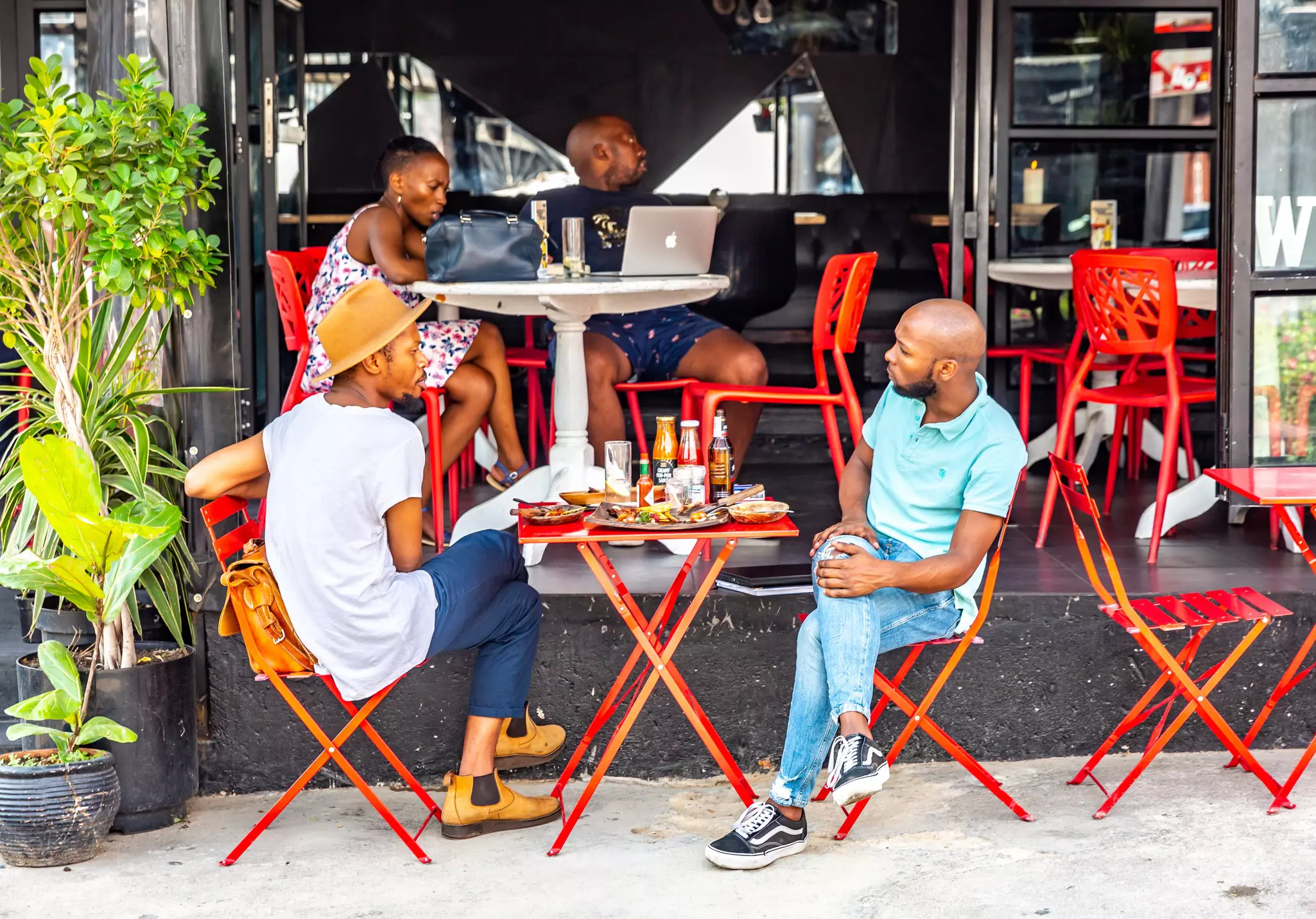 Two diners sit at a red metal table on a sidewalk in front of a cafe.