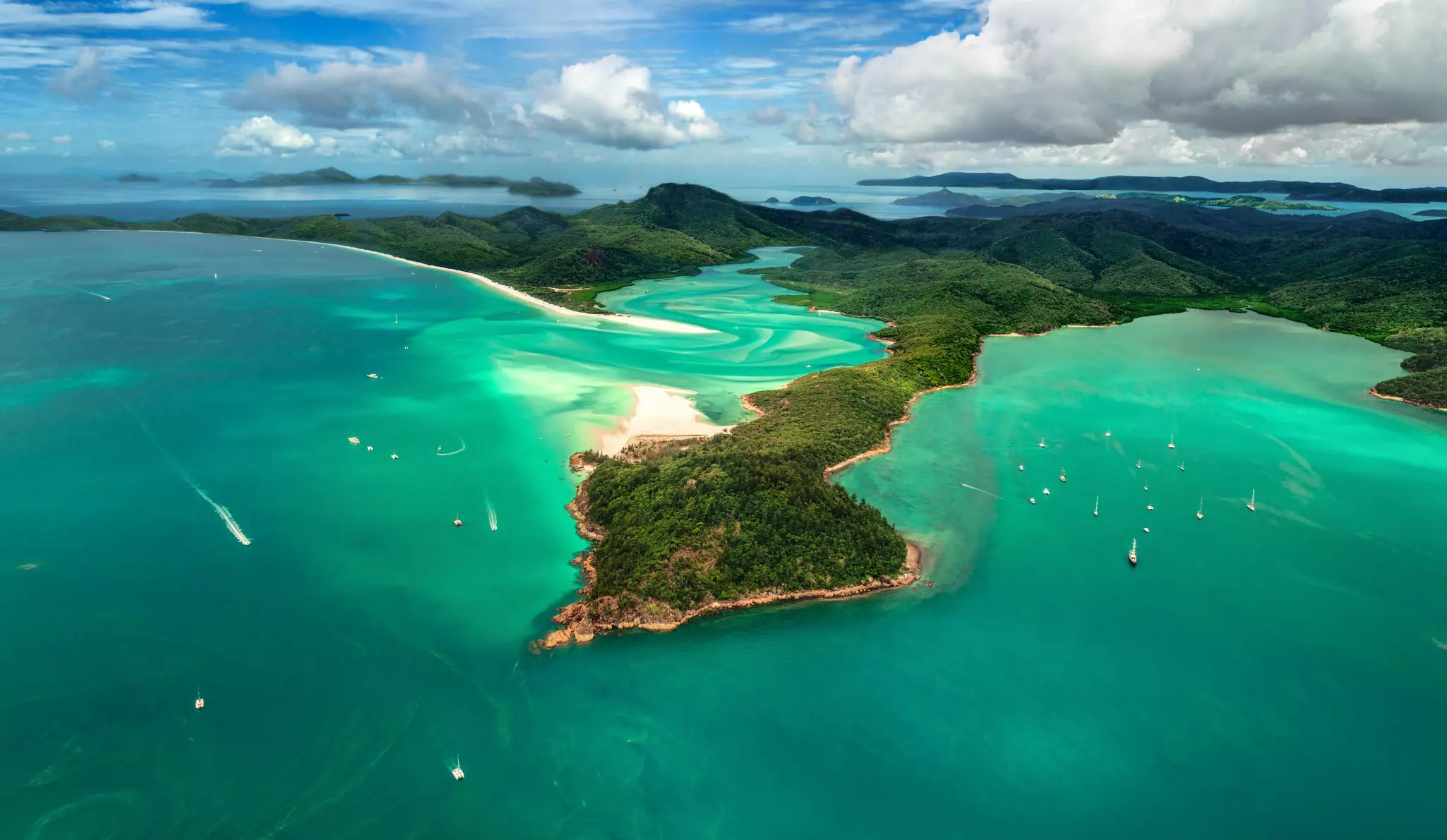 A lush island surrounded by turquoise water, with smaller islands on the horizon; boats are anchored in coves, and the wakes of speedboats leave white streaks in the water.