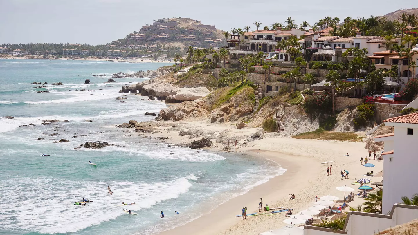 A wide view of a cliffs leading down to a beach lapped by white surf. People are on the beach and villas sit at the top of the cliffs.