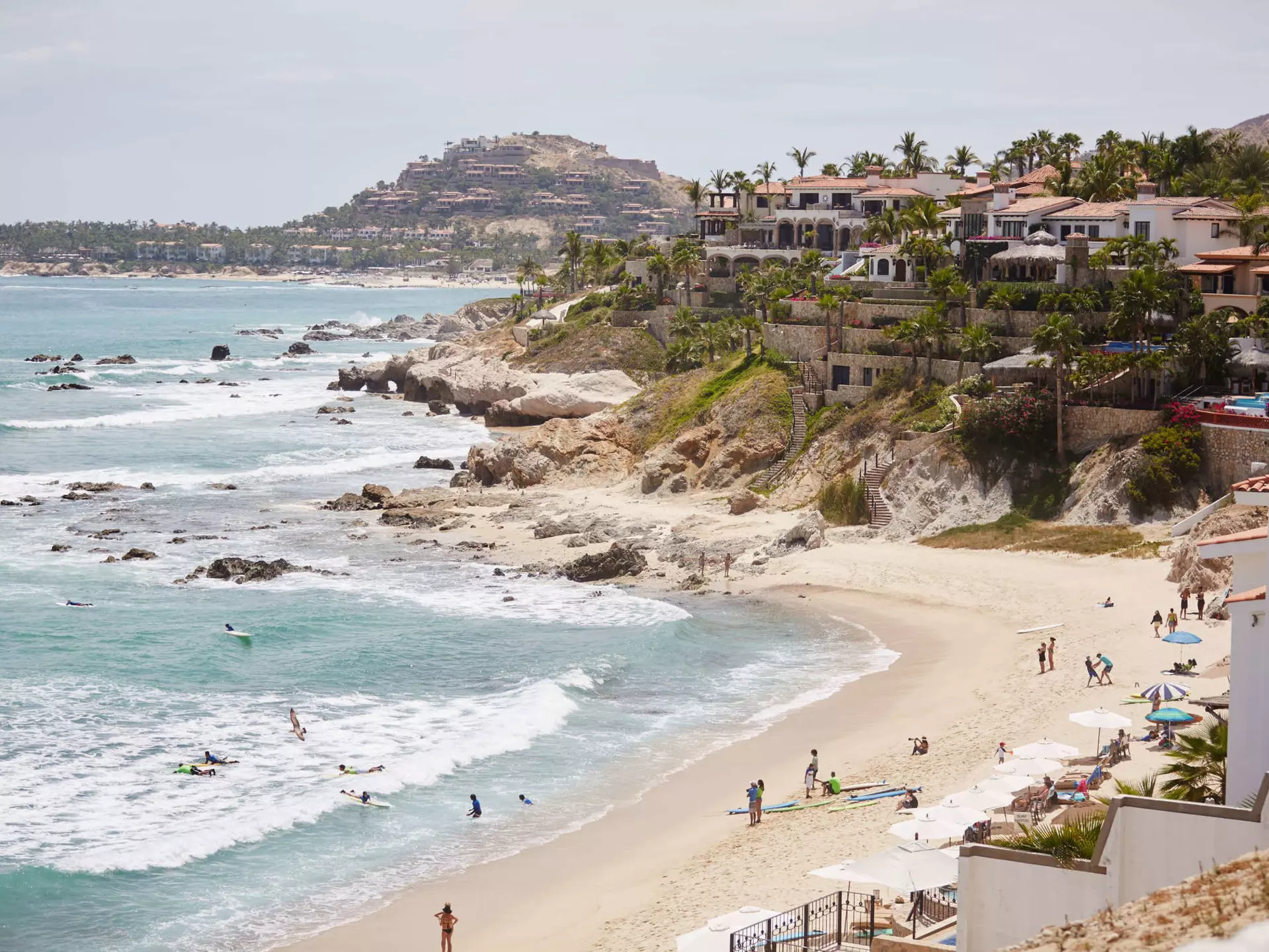 A wide view of a cliffs leading down to a beach lapped by white surf. People are on the beach and villas sit at the top of the cliffs.