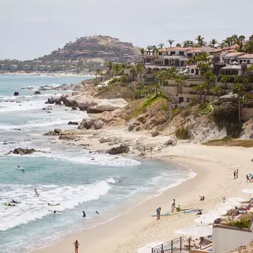 A wide view of a cliffs leading down to a beach lapped by white surf. People are on the beach and villas sit at the top of the cliffs.