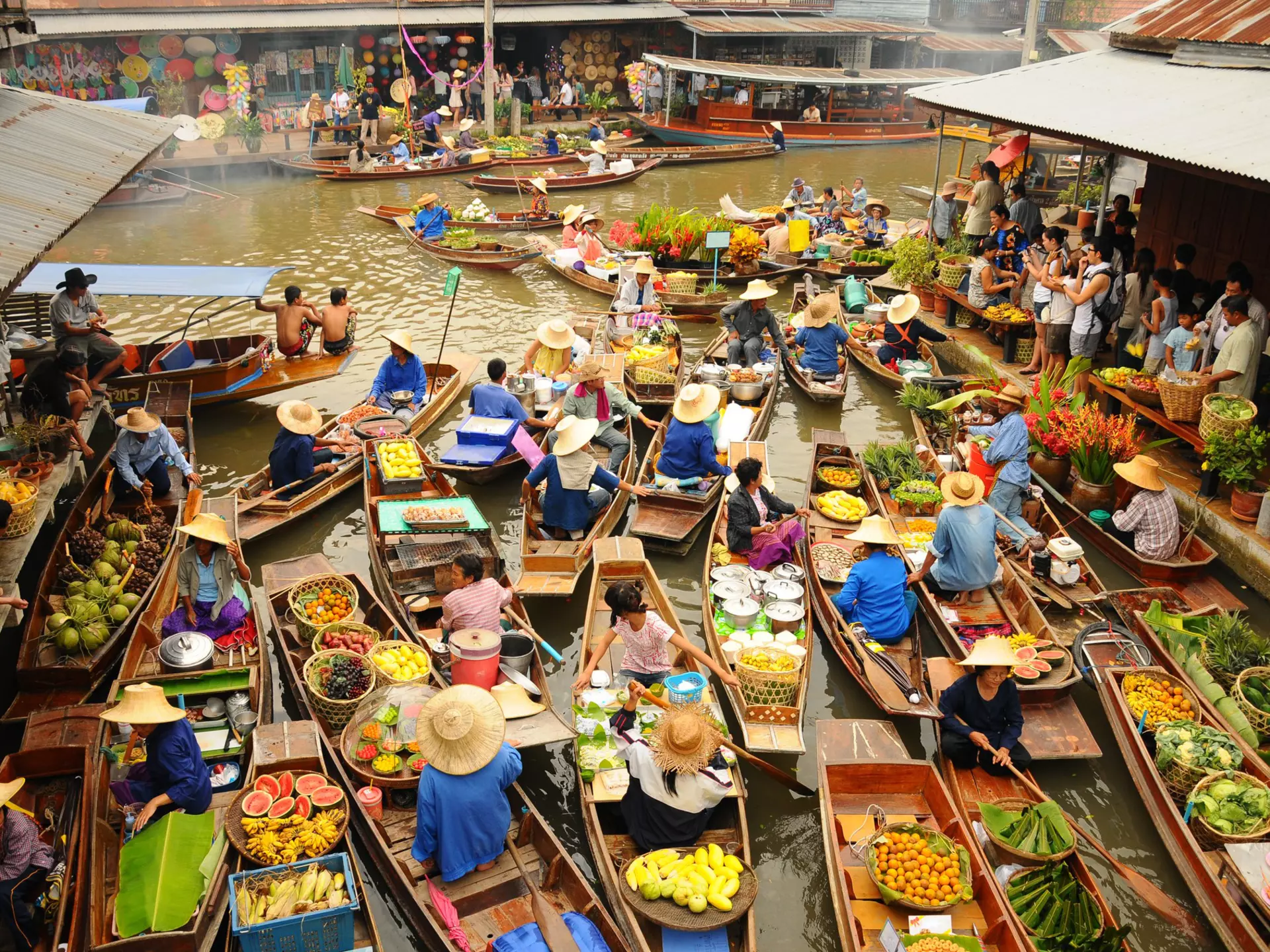 A vast amounts of small wooden boats gather to trade goods at  Amphawa floating market. Most of the boats sell food, specifically fruits. Tourist wielding cameras watch on.
