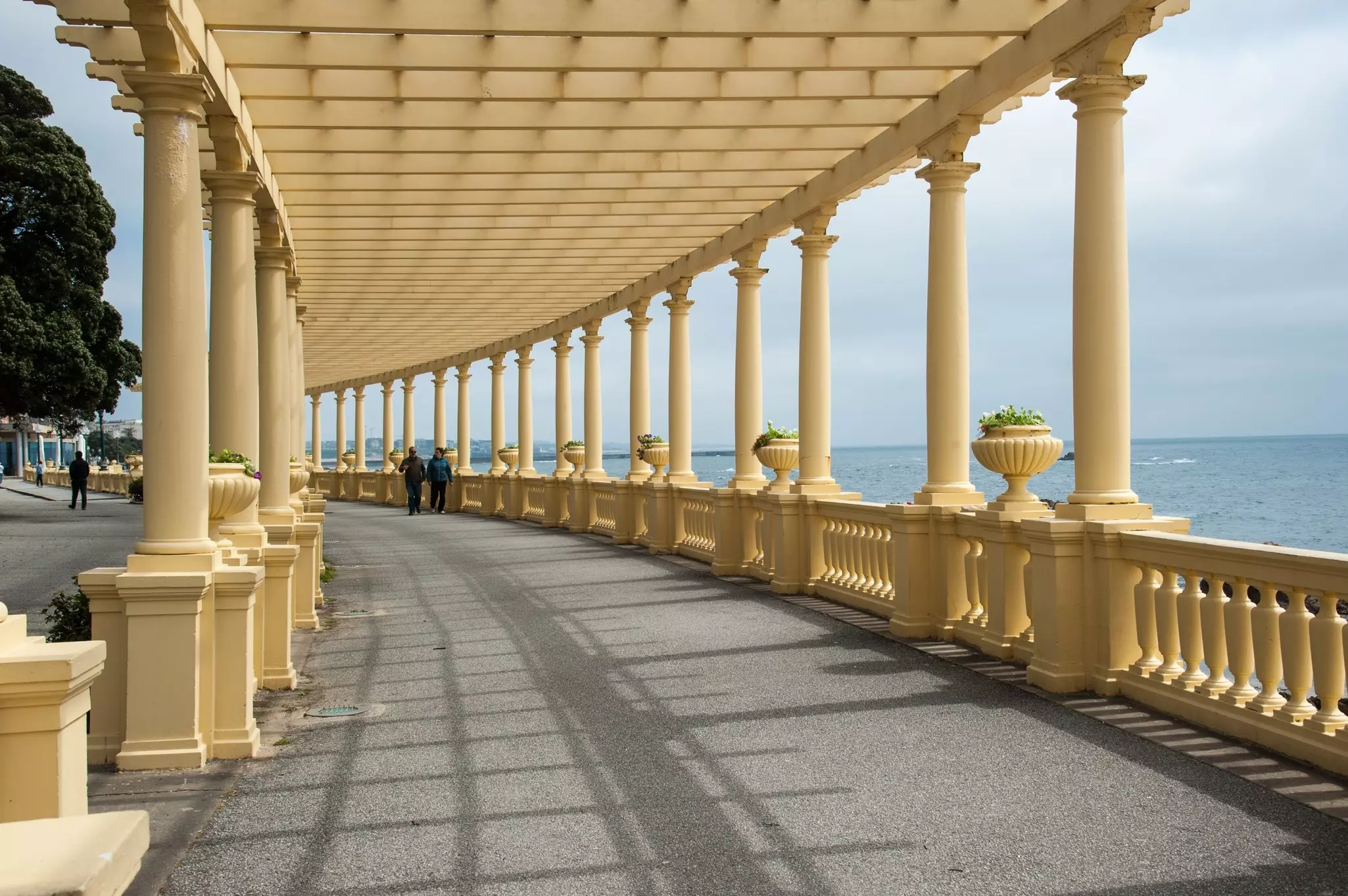 Two people walking down a long curving white portico by the ocean.