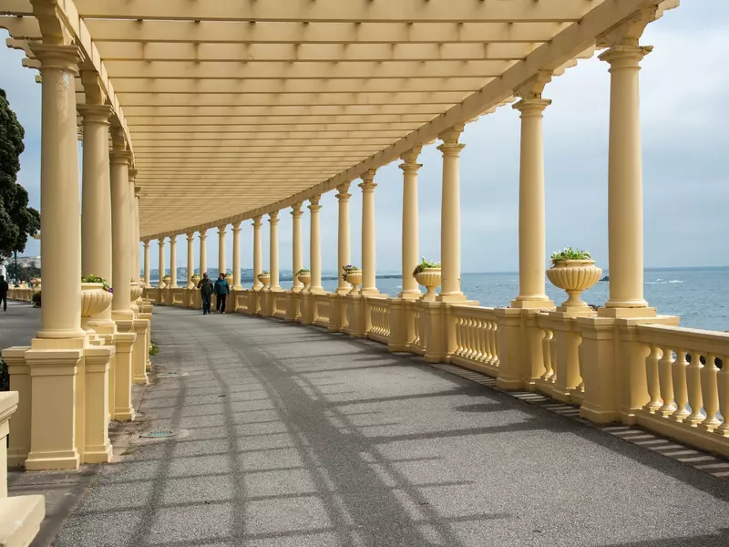 Two people walking down a long curving white portico by the ocean.