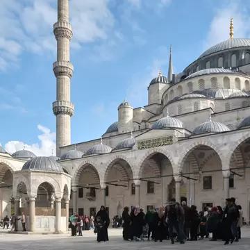 Blue Mosque courtyard in Sultanahmet with a domed ablution fountain, minaret and arcaded portico; visitors on marble paving