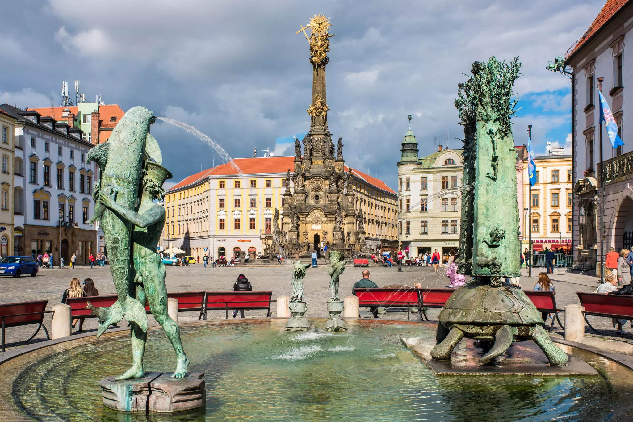An elaborately carved column is seen beyond two sculptural fountains in a city square