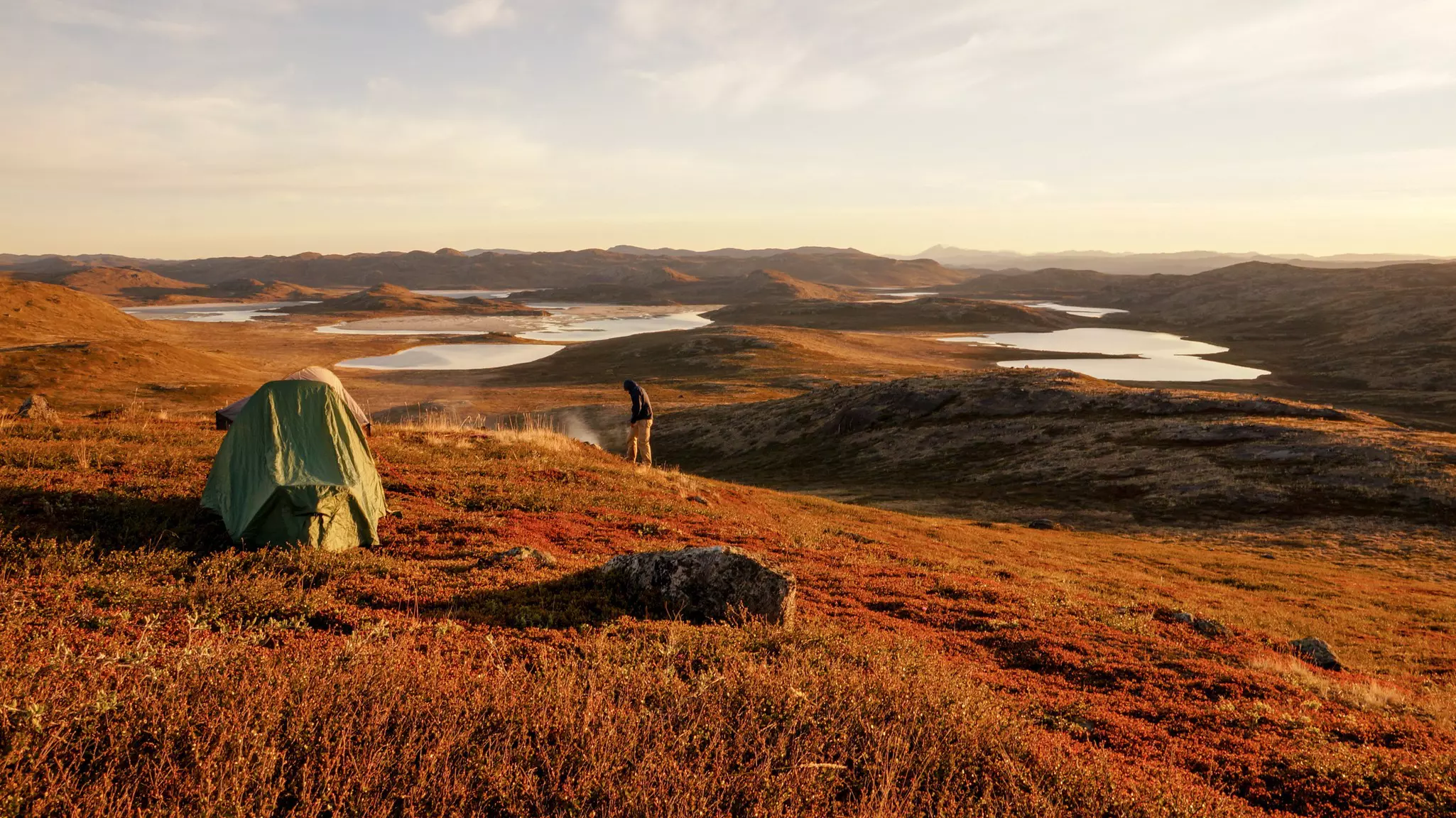 A person stands near a small green tent in a landscape lit by warm light; the vegetation is orangish brown, and there are pools of water and hills in the background.