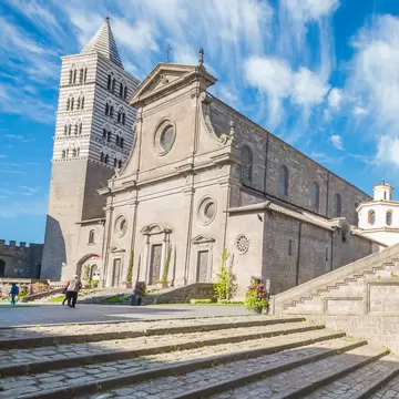 Stone cathedral with a striped bell tower beside a grand outdoor staircase and arched bridge, cobbled steps under blue sky.