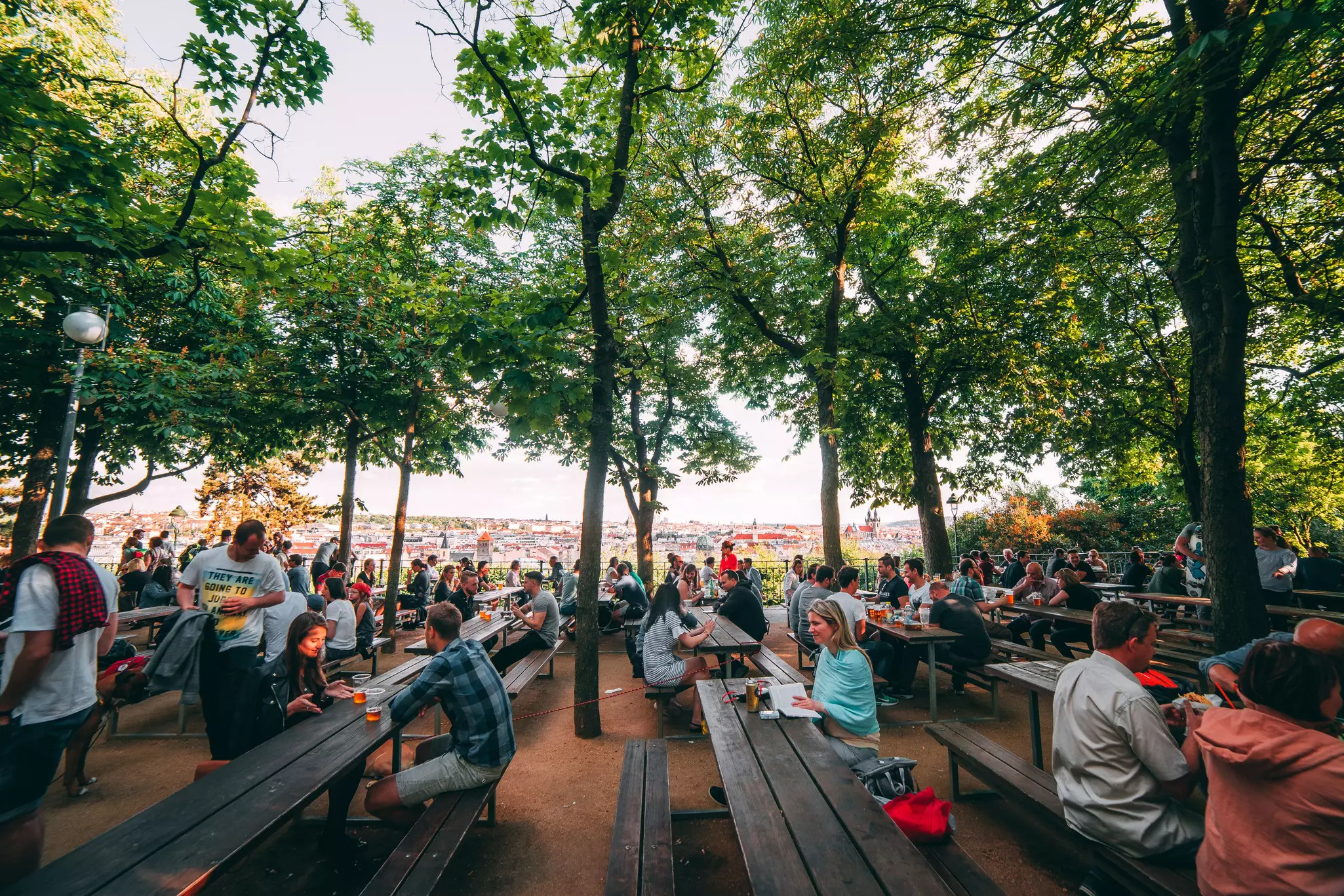 People sitting at long wooden picnic tables under trees in Prague during the summer.