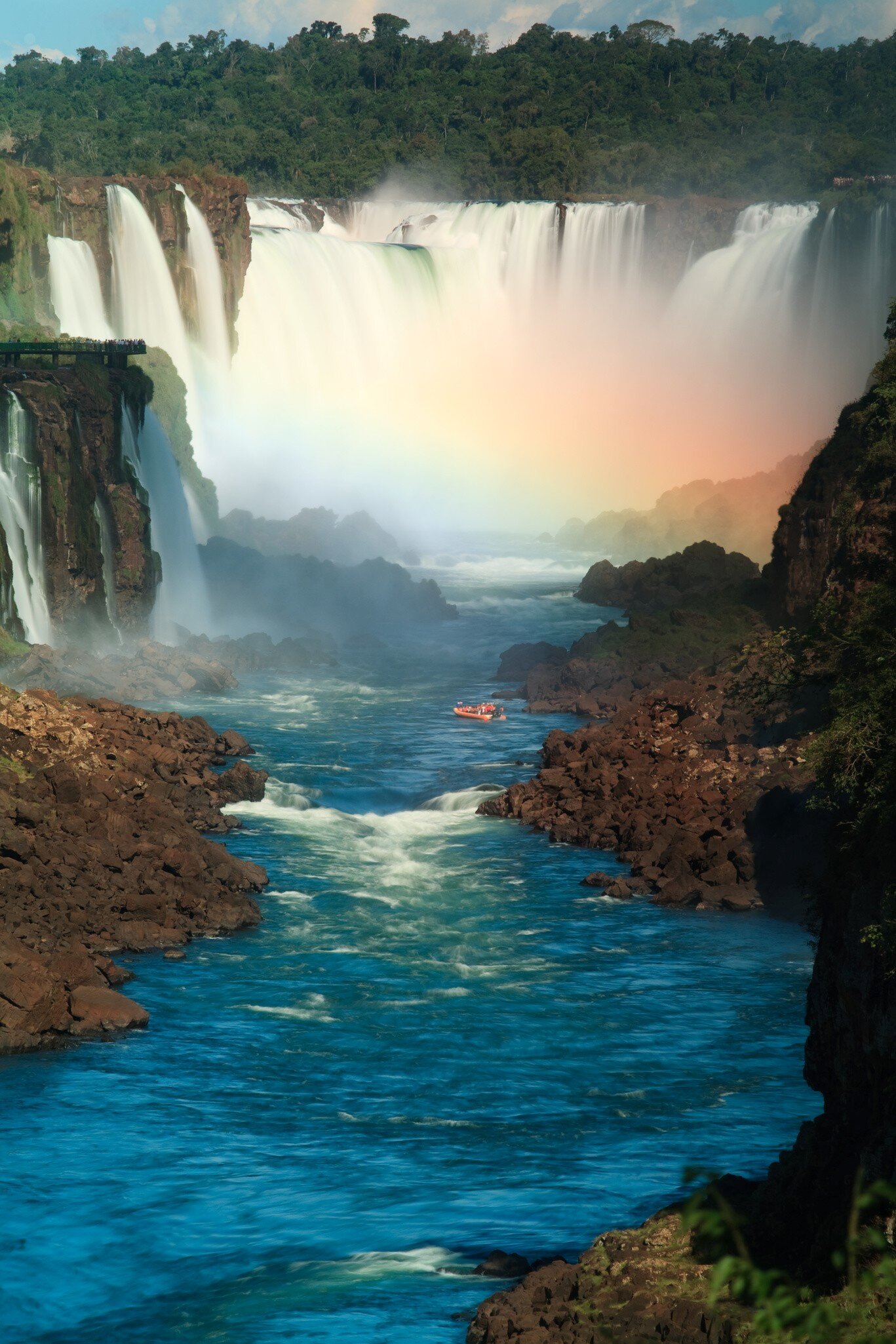 Wide curtains of waterfalls pouring into a misty gorge with a rainbow, a red boat on the blue river between rocky banks