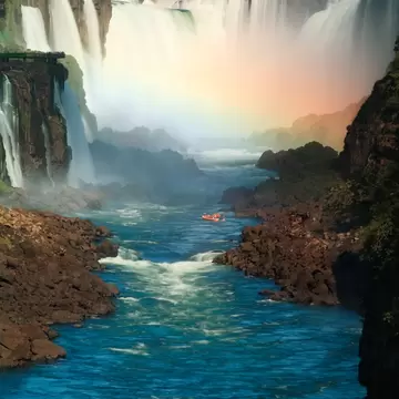 Wide curtains of waterfalls pouring into a misty gorge with a rainbow, a red boat on the blue river between rocky banks