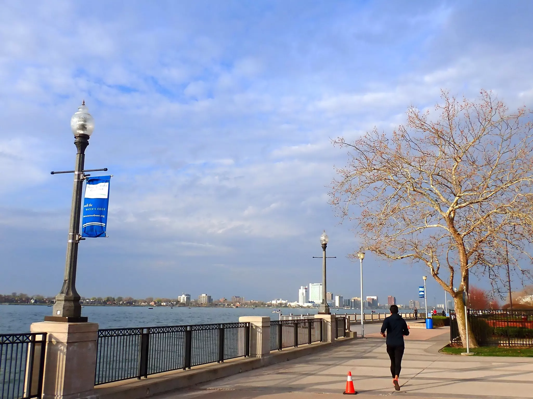Person jogging along a cement riverfront path with city skyline in the distance on a mostly sunny day.