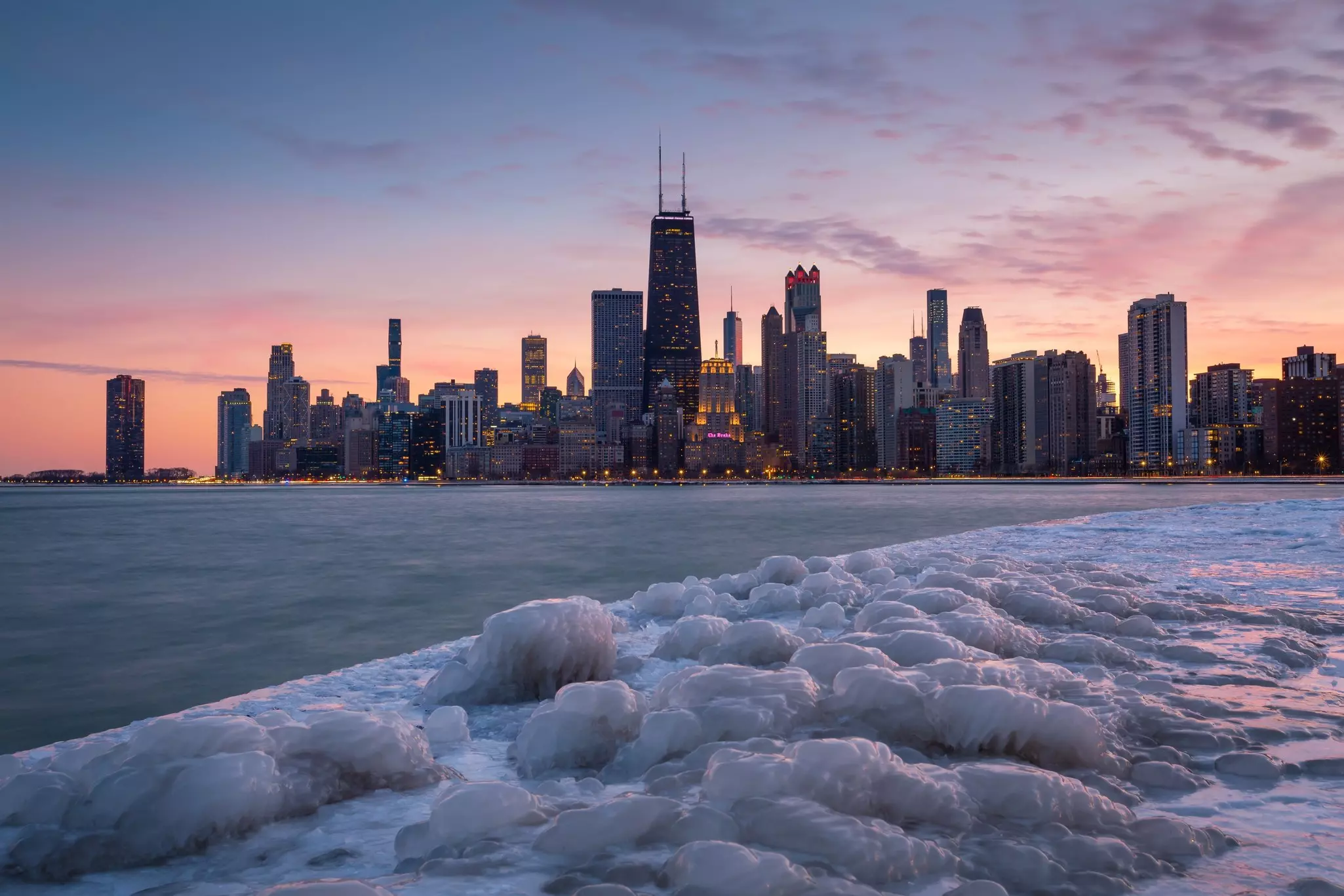 Ice on the surface of a frozen lake is seen in front of a city skyline silhouetted by dusk light.