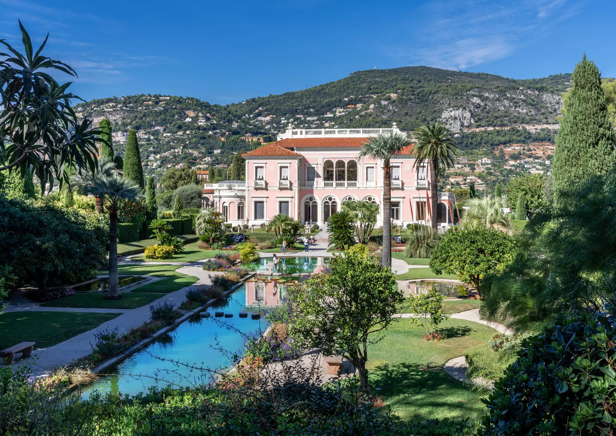 A grand pastel-pink villa in landscaped gardens. Visitors wander around.