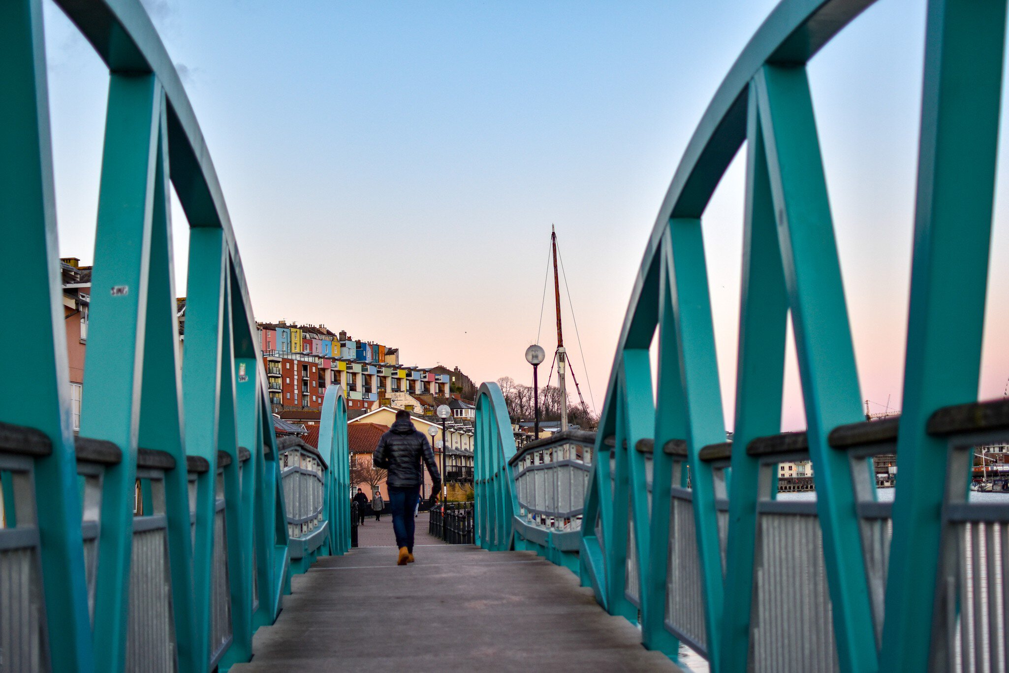 Man walking on Blue / Turquoise footbridge leading from Bristol Harbor towards the well known colorful houses of Clifton -Image; Shutterstock ID 1330383716; your: Sloane Tucker; gl: 65050; netsuite: Online Editorial; full: Destination Page