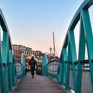 Man walking on Blue / Turquoise footbridge leading from Bristol Harbor towards the well known colorful houses of Clifton -Image; Shutterstock ID 1330383716; your: Sloane Tucker; gl: 65050; netsuite: Online Editorial; full: Destination Page