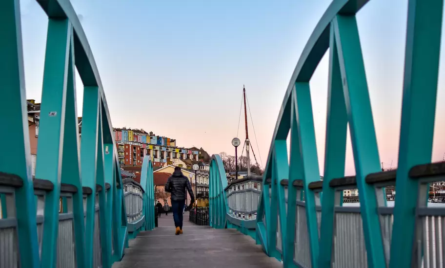 Man walking on Blue / Turquoise footbridge leading from Bristol Harbor towards the well known colorful houses of Clifton -Image; Shutterstock ID 1330383716; your: Sloane Tucker; gl: 65050; netsuite: Online Editorial; full: Destination Page